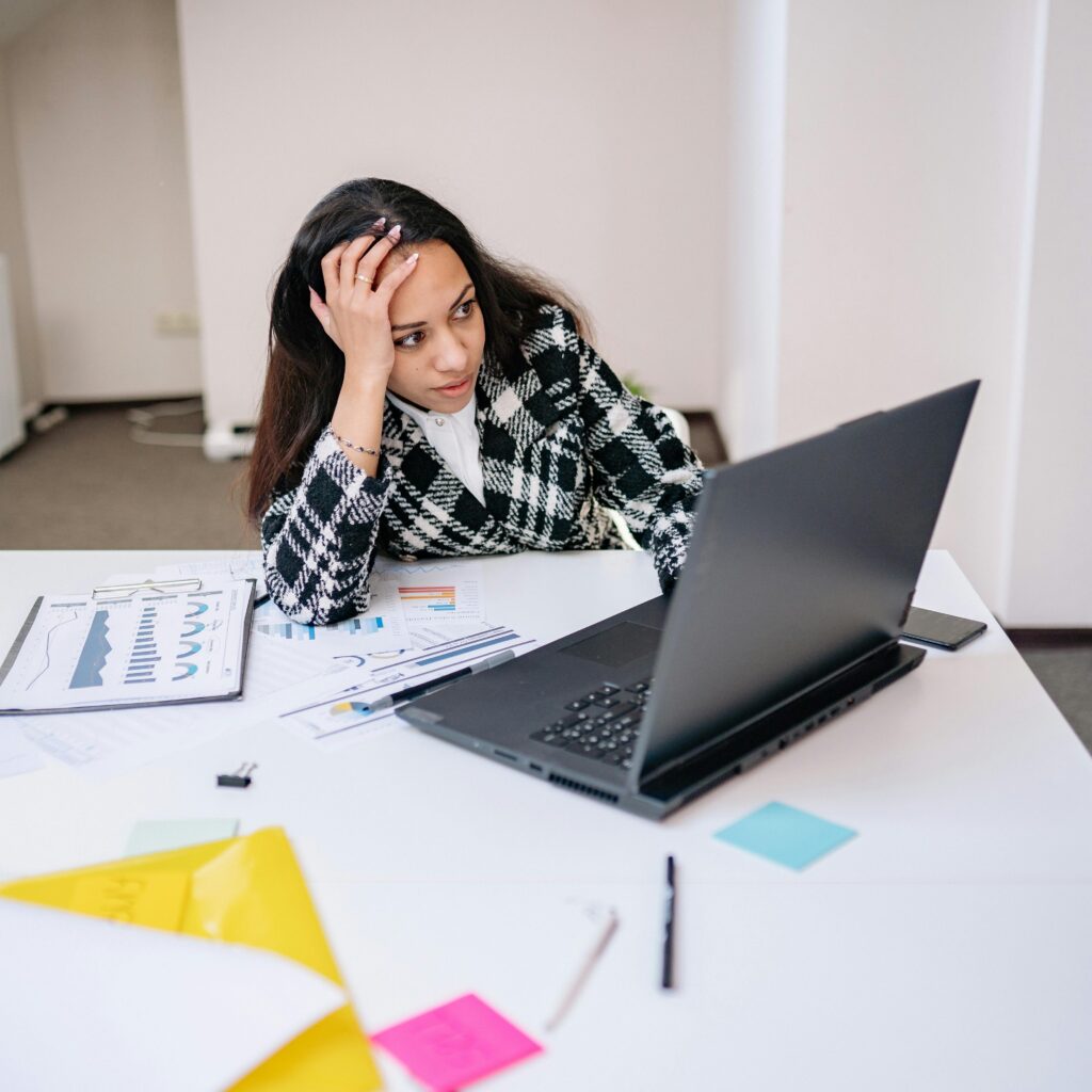 A frustrated woman sits at a desk overwhelmed with work tasks on her laptop.